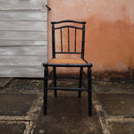 A black wooden chair with a woven cane seat, positioned in front of a textured peach-colored wall and a weathered gray wooden door.