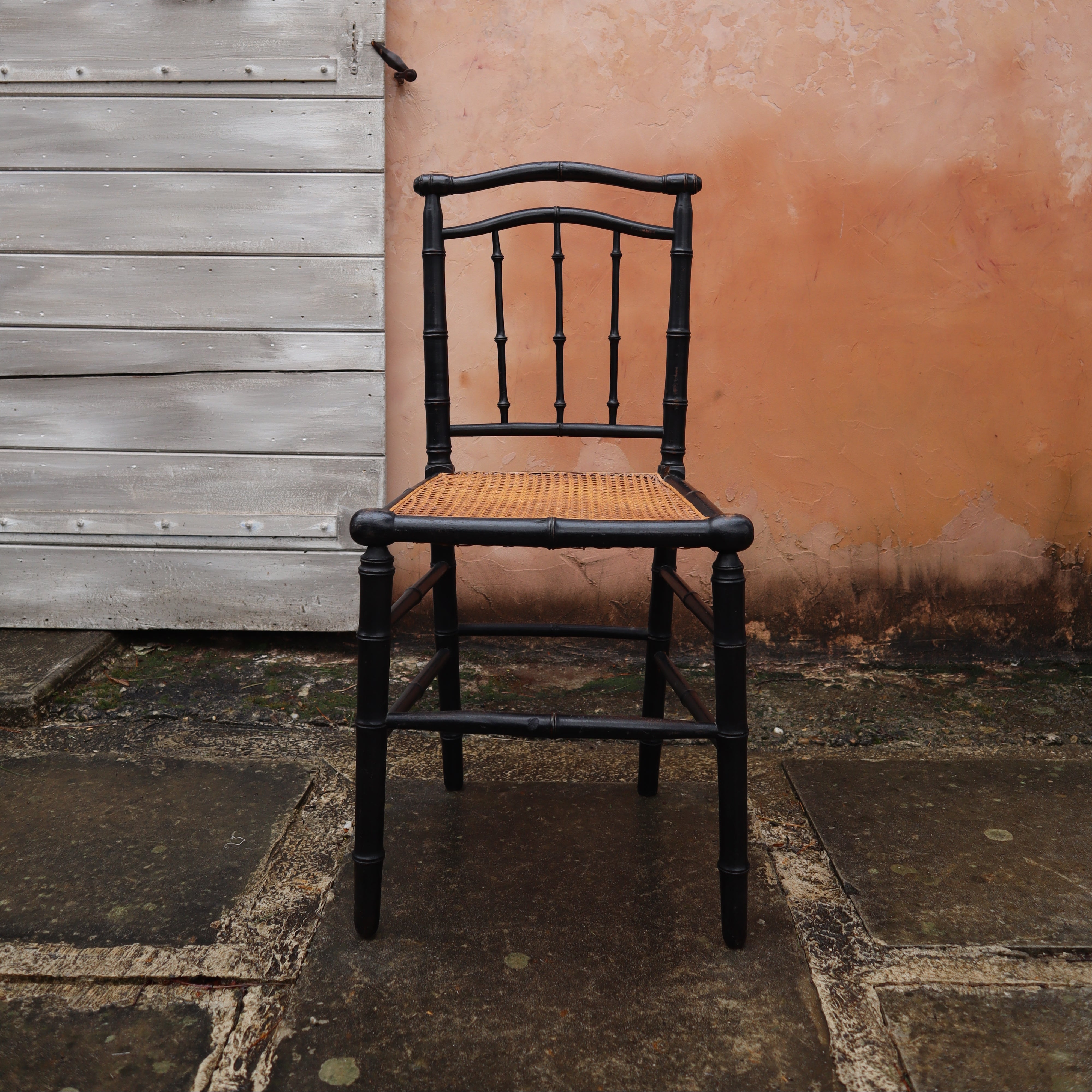 A black wooden chair with a woven cane seat, positioned in front of a textured peach-colored wall and a weathered gray wooden door.