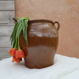 A rustic brown ceramic clay pot with two handles sits on a white fabric-covered surface, with orange tulips draped over the left side against a peach-colored wall and light wood panel background.