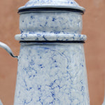 A close-up of a vintage enamel coffee pot or teapot with a blue and white marbled pattern, featuring a lid with a small spout on the left, set against a solid terracotta-colored background.