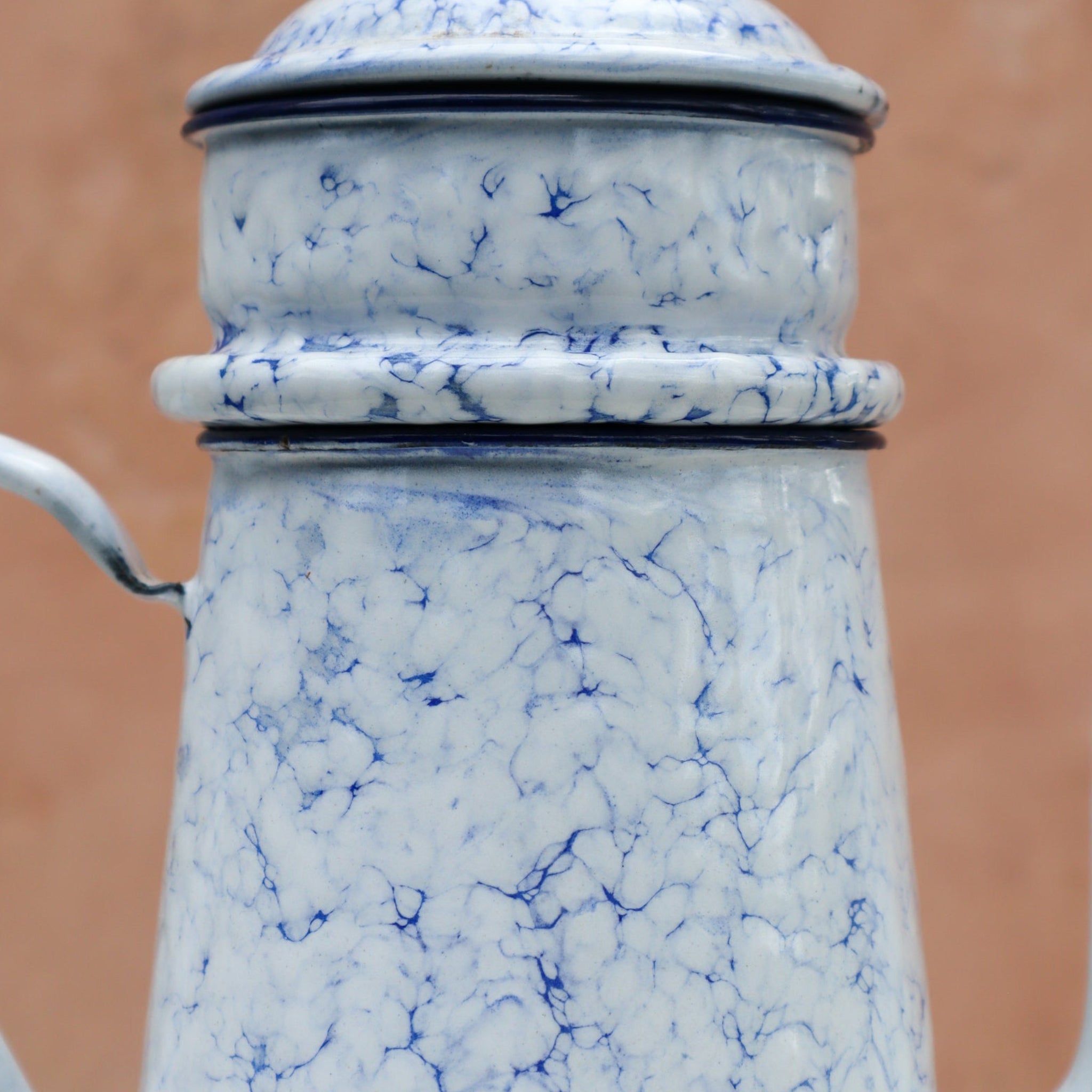 A close-up of a vintage enamel coffee pot or teapot with a blue and white marbled pattern, featuring a lid with a small spout on the left, set against a solid terracotta-colored background.