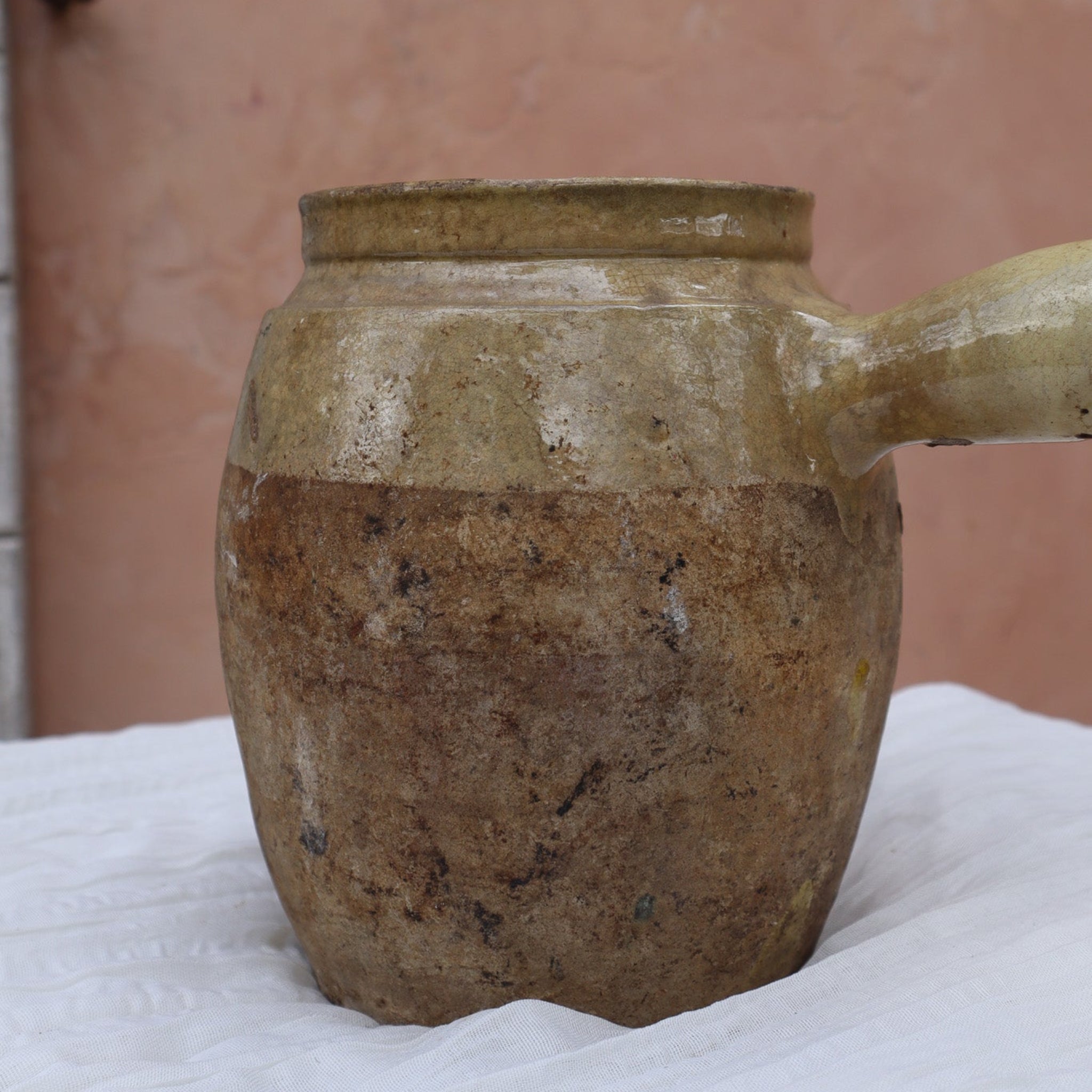Brown ceramic pot with a handle on a white surface against a pinkish-brown background