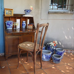 A styled interior scene with a wooden writing desk, two wooden chairs, and multiple blue-and-white ceramic vases and flower pots on terracotta tile flooring against a beige wall and blue door.