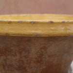 Close-up of a rustic ceramic pot or planter with a yellow glazed rim and a brown textured body, set against a soft-focus terracotta background.