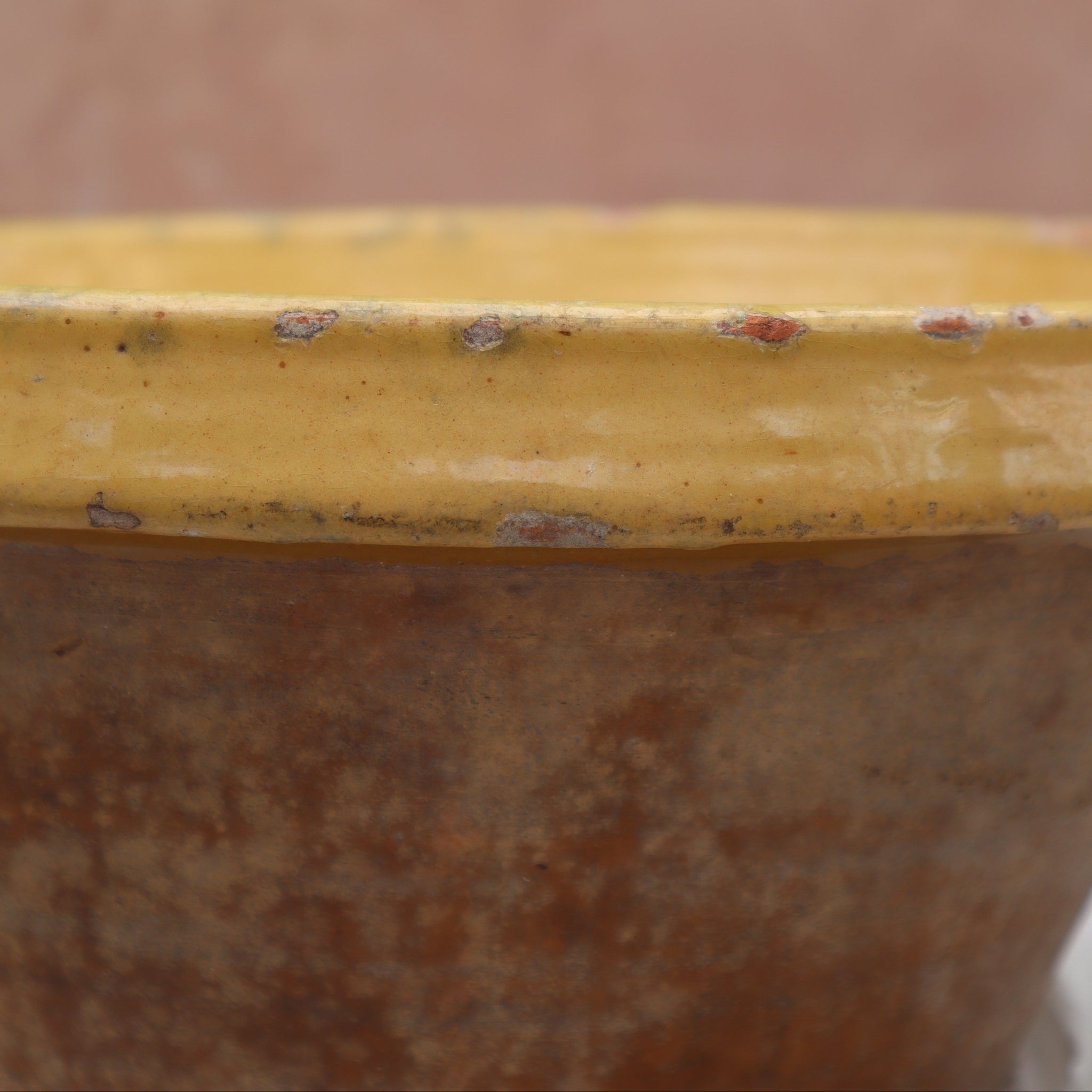 Close-up of a rustic ceramic pot or planter with a yellow glazed rim and a brown textured body, set against a soft-focus terracotta background.