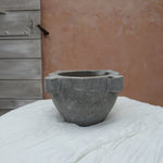 A gray stone mortar and pestle set on a white fabric-covered surface, with a rustic terracotta wall and weathered wooden door in the background.