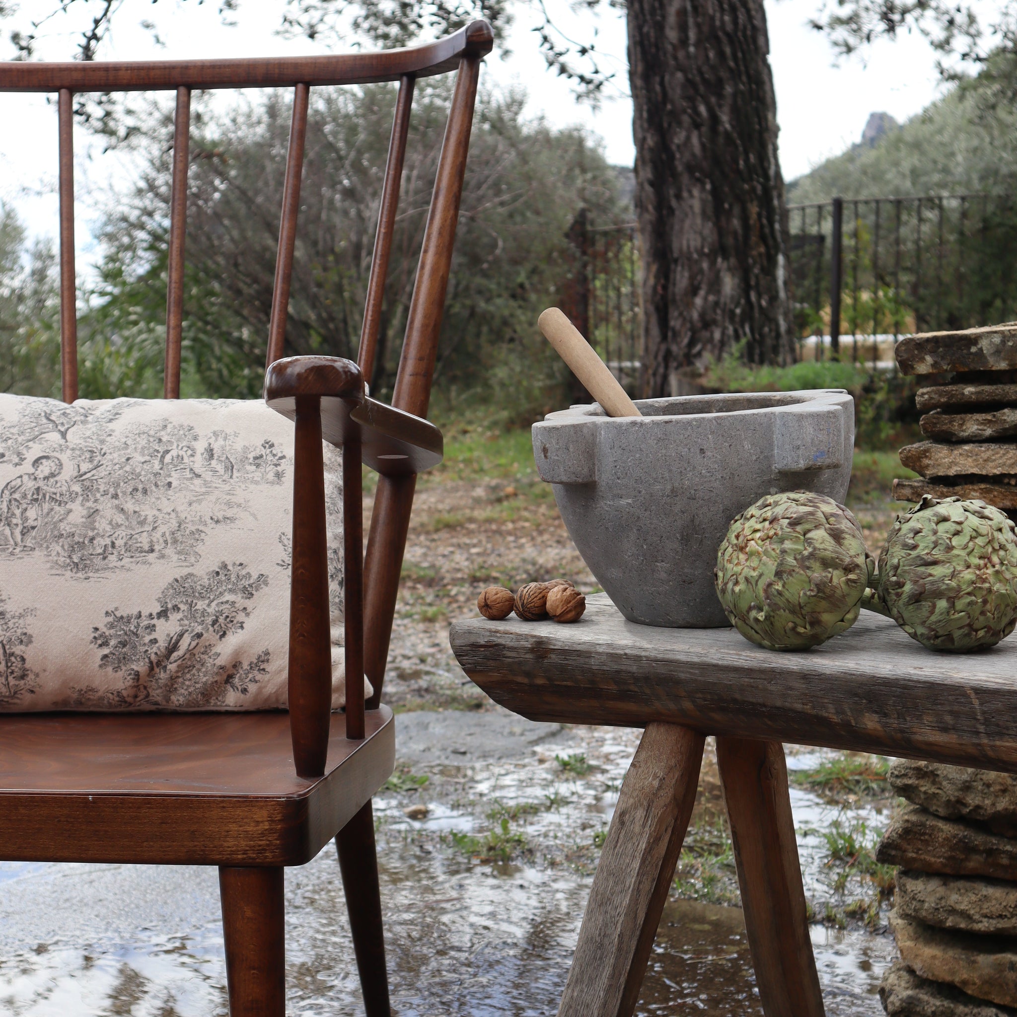 A rustic outdoor scene featuring a dark brown wooden chair with a beige toile-patterned cushion, next to a weathered wooden table holding a gray stone mortar and pestle, two green artichokes, and walnuts on a wet stone-paved surface.