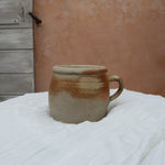 A handmade ceramic mug with a rustic, earthy glaze sits on a white fabric-covered table. The background features a textured terracotta plaster wall and a weathered wooden door.