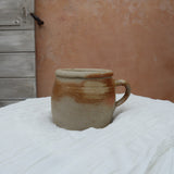 A handmade ceramic mug with a rustic, earthy glaze sits on a white fabric-covered table. The background features a textured terracotta plaster wall and a weathered wooden door.