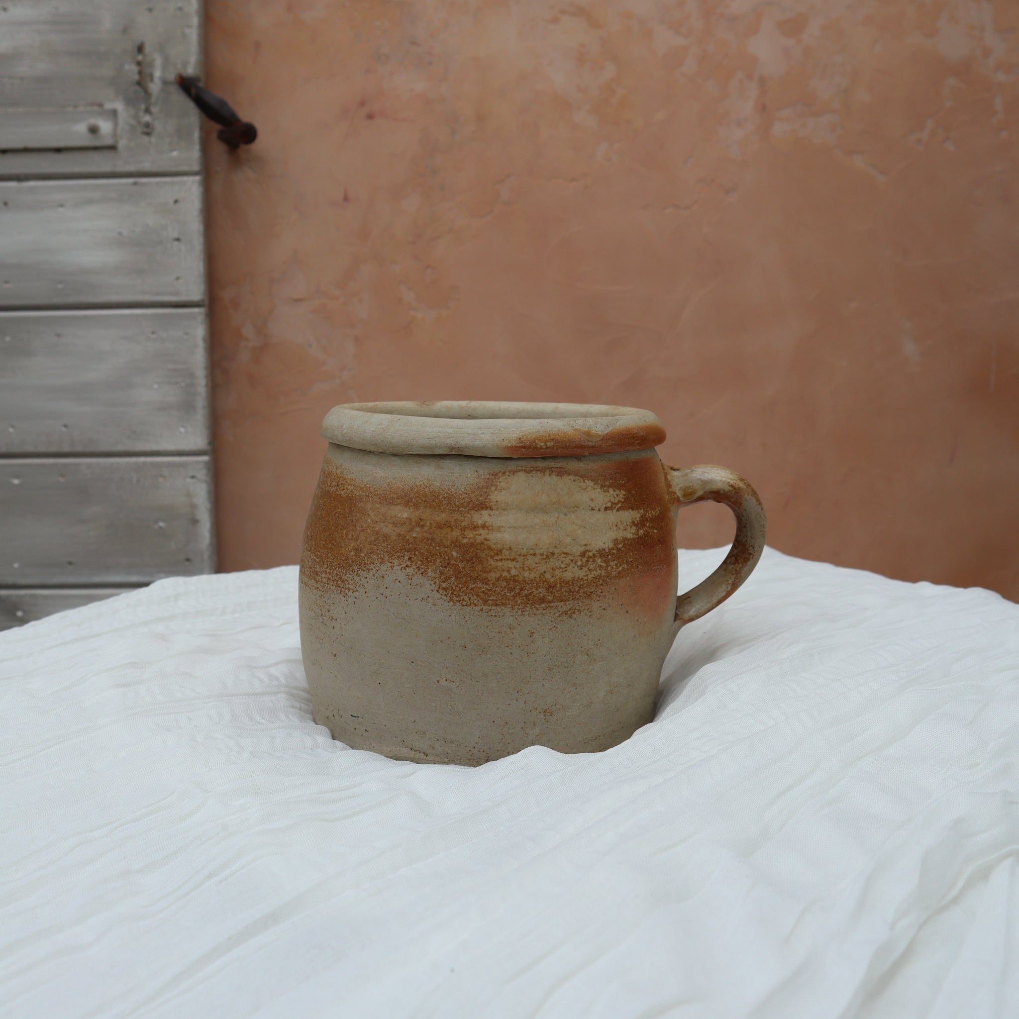 A handmade ceramic mug with a rustic, earthy glaze sits on a white fabric-covered table. The background features a textured terracotta plaster wall and a weathered wooden door.