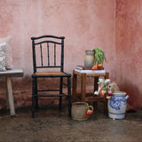 A rustic interior corner with a pink plaster wall, a black spindle-back chair, a small wooden side table with books and a vase of flowers, and a blue-and-white ceramic pot on the floor.