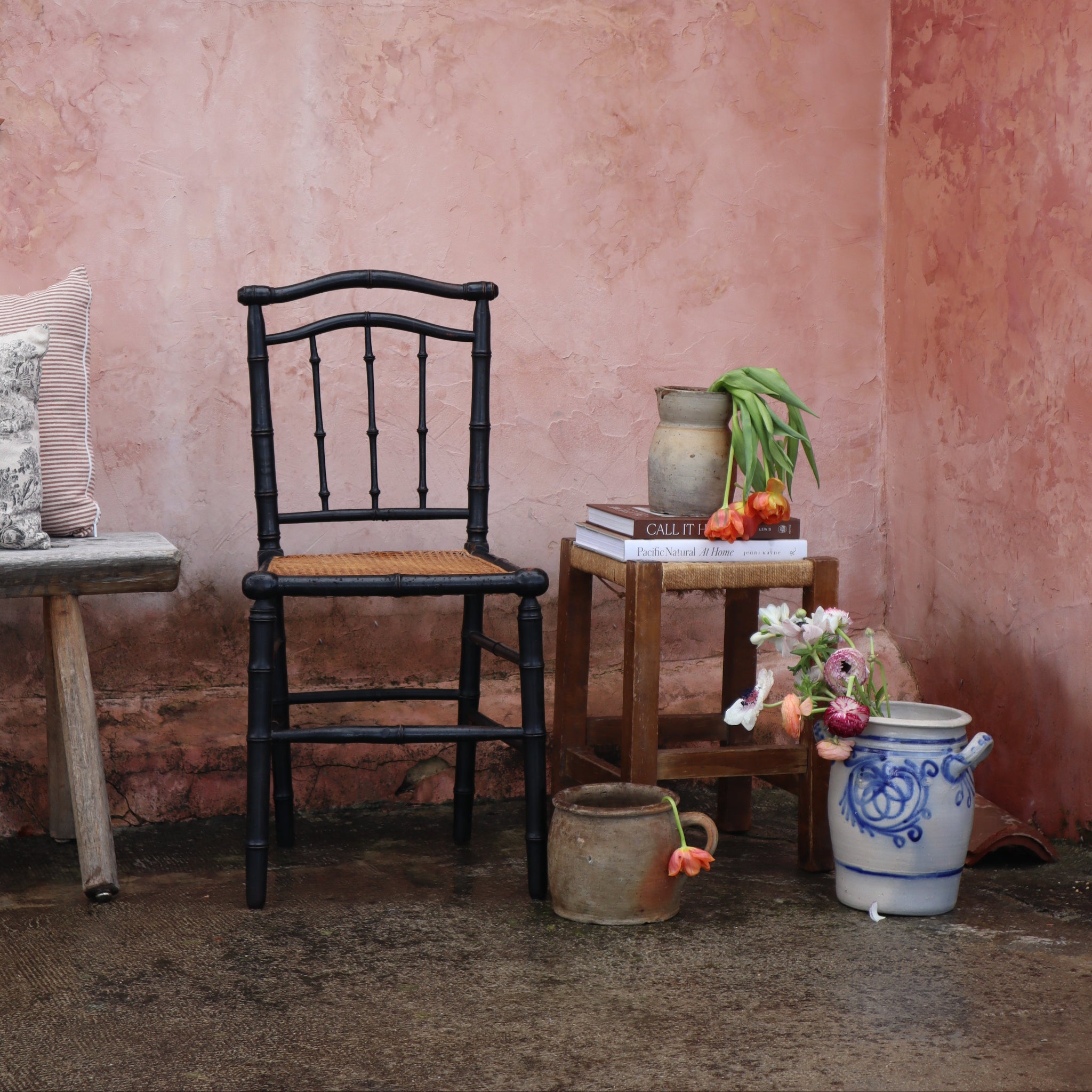 A rustic interior corner with a pink plaster wall, a black spindle-back chair, a small wooden side table with books and a vase of flowers, and a blue-and-white ceramic pot on the floor.